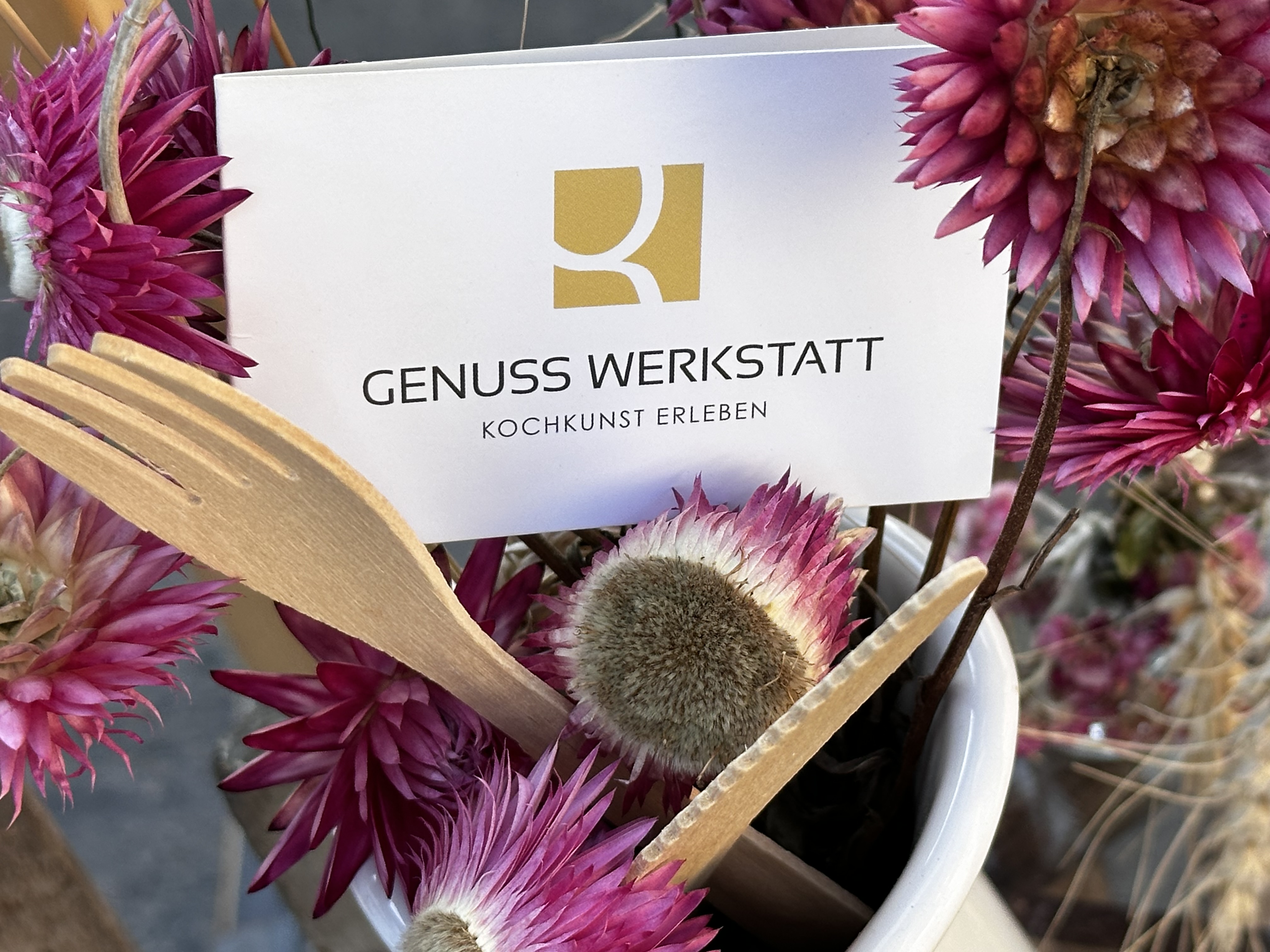 Genuss-Werkstatt-Karte zwischen Strohblumen und Holz-Besteck — Branding-Detail vor Ort